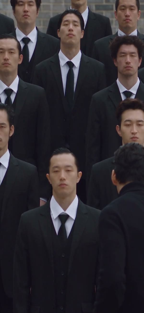 Sunglasses-wearing lead in front of a suited crowd